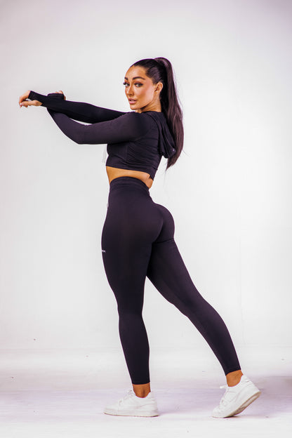 A woman wearing a black coloured gym high waisted leggings with a matching long sleeve cropped top with hoodie